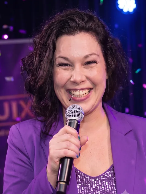 Presentatrice Roxy in paars colbert Presentatrice Roxy met donkerbruin krullend haar in een paars colbert en pailletten top spreekt door een microfoon op een QuizX-avond.